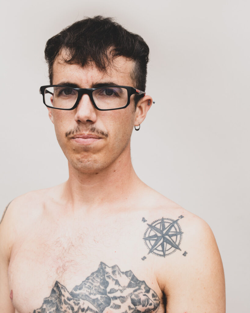 Person med briller og overskæg står i bar overkrop med tatoveringer og kigger direkte i kameraet.