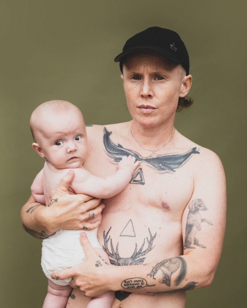 Person med bar overkrop og tatoveringer holder et lille barn i favnen