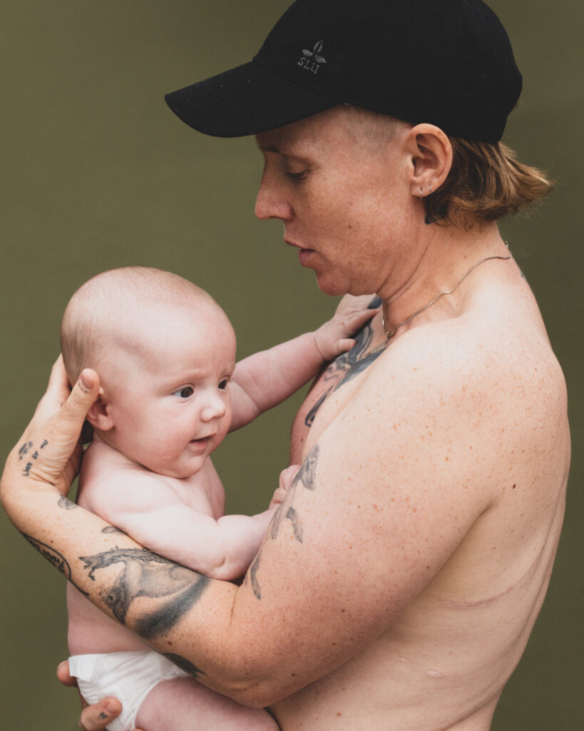 Person med kasket og bar overkrop ses i profil holde et spædbarn.
